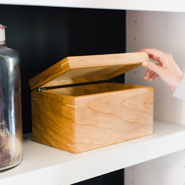 a woman's hand opening a cherry keepsake box sitting on a bookshelf, with hard maple corner spline joinery