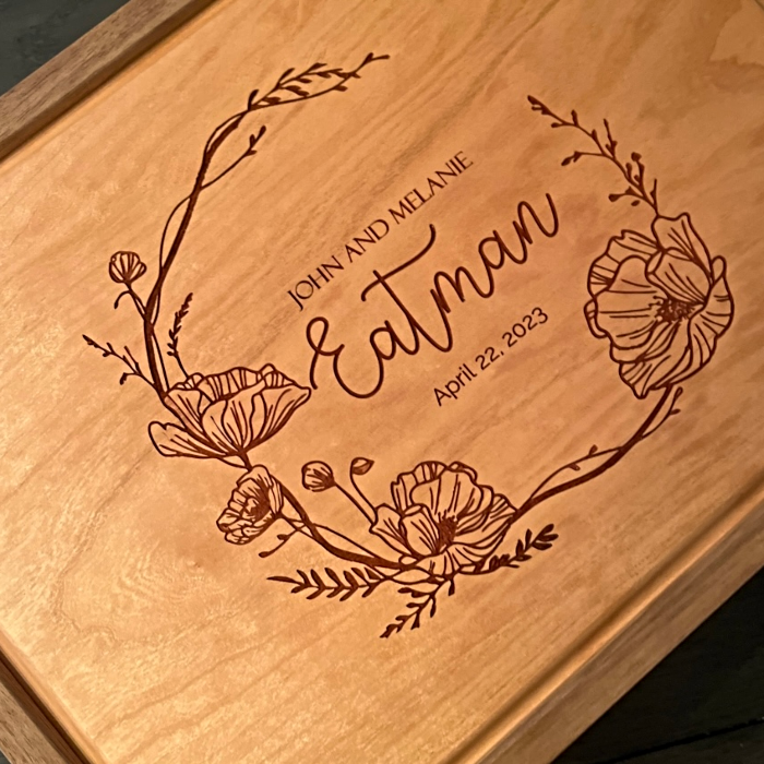 Laser-engraved floral wreath design with the names "John and Melanie Eatman" and date "April 22, 2023" on a cherry wood box lid; an example of personalized engraving available from Mad Tree Woodcrafts.