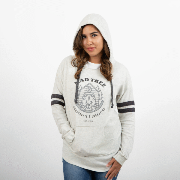 Woman wearing a light gray Mad Tree Woodcrafts & Engraving hoodie with the hood up, one hand in the front pocket and the other resting at her side, looking forward with a subtle smile.