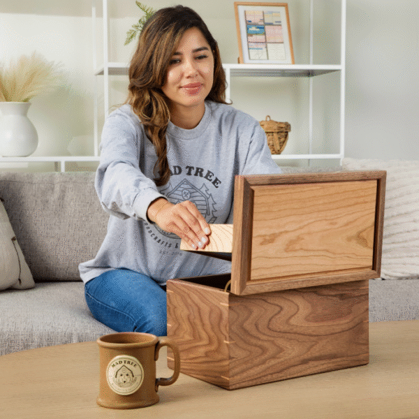 Woman seated on a gray couch, lifting the removable tray from a large walnut keepsake box; a brown ceramic Mad Tree Woodcrafts coffee mug rests on the wooden coffee table in front of her.