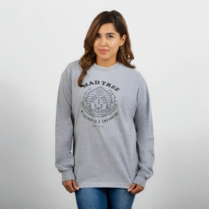 Woman wearing a heather gray long-sleeve Mad Tree Woodcrafts & Engraving shirt, standing with one hand holding the hem and looking slightly to the side with a subtle smile.