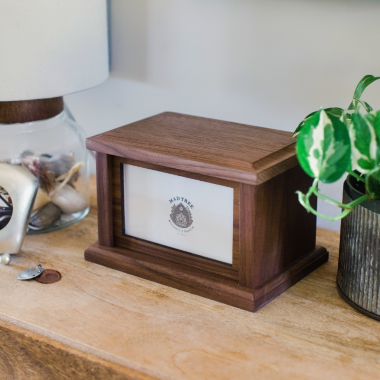 Handcrafted walnut keepsake box with framed front panel, displayed on a wooden table beside a potted plant and decorative lamp.