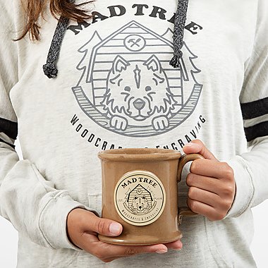 Person holding a brown stoneware Mad Tree mug in front of a Mad Tree hoodie logo