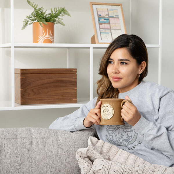 Woman sitting on a couch holding a Mad Tree Woodcrafts mug, with a walnut keepsake box on the shelf behind her.