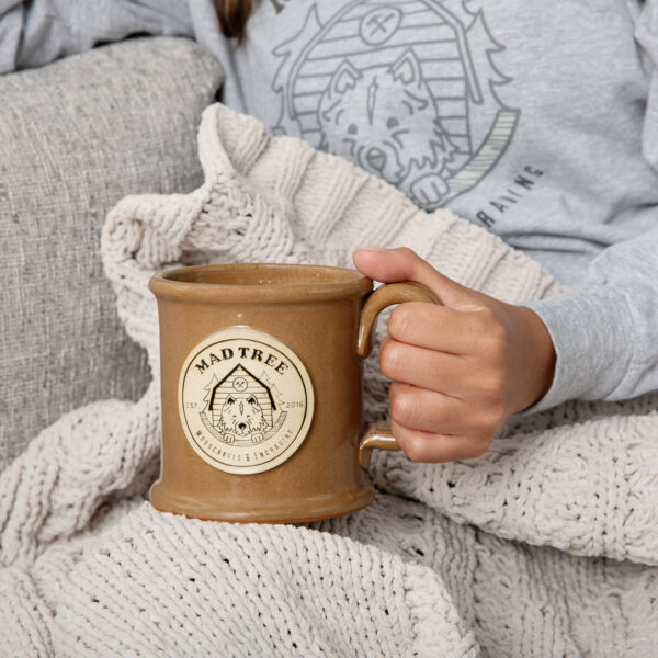 Hand holding a brown stoneware Mad Tree mug with a raised logo medallion, resting on a cozy knit blanket with a Mad Tree hoodie in the background.