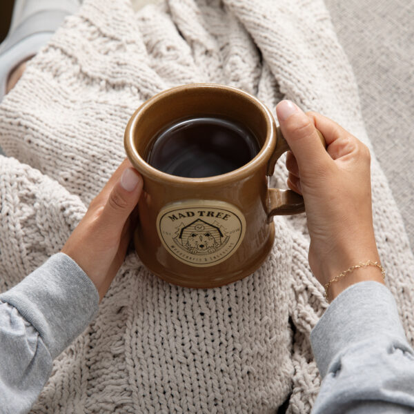 Close-up of hands holding a brown Mad Tree Woodcrafts & Engraving ceramic mug filled with coffee, resting on a soft beige knitted blanket.
